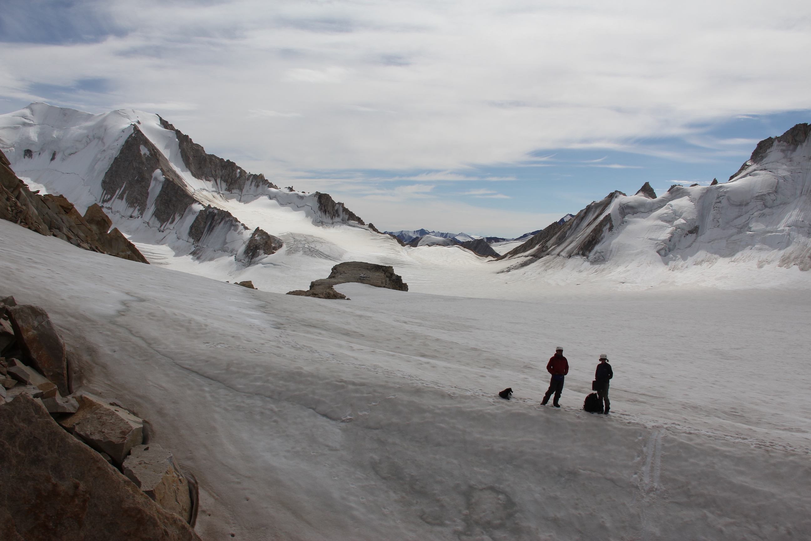 Scenic shot of mountains in background. Two people in the foreground stand on a vast expanse of ice, looking towards the mountains.